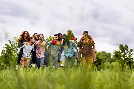 Group of multiethnic young cheerful people walking together and playing piggyback in the countryside - happy friends having fun gathering in the weekend - trust, unity and diversity lifestyle concept