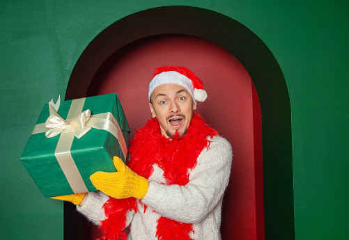 Young handsome happy surprised man wearing Santa hat holding Christmas gift box on the red arch background. New Year style