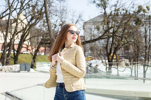 A beautiful blonde girl in a short light jacket and sunglasses walks around the city, enjoying the spring weather.