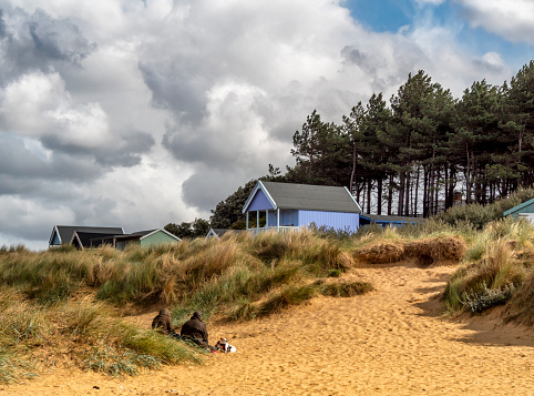 Wooden beach huts in shades of blue and green nestling amongst the sand dunes at Old Hunstanton in North West Norfolk. Two people and a dog take shelter from the stiff breeze.