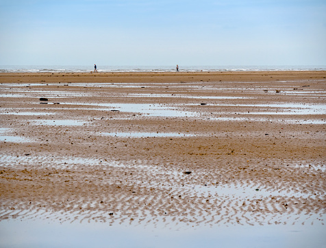 Two people stroll by the water’s edge on the vast beach at Old Hunstanton in North West Norfolk, Eastern England, at low tide. Seashells and seaweed litter the shore and the sand has formed into ripples by the action of the waves.