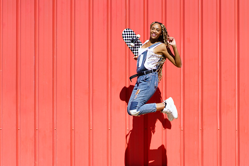 Black young woman wtih a skateboard jumping with happiness on red urban wall background.