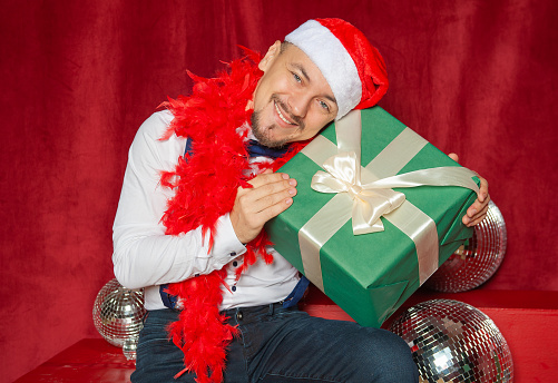 Young handsome smiling man wearing Santa hat with Christmas gift box sitting on the red background. New Year style
