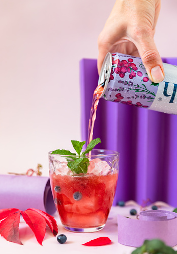 Kaliningrad, Russia - October 19, 2022: Female hand pouring pink grape soda drink from aluminum can to glass with ice cubes and mint, pink background. Carbonated non-alcoholic drink.