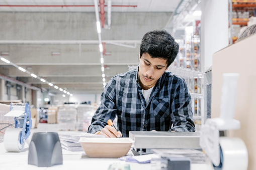 Young male employee working at a desk in distribution warehouse. Man in casuals wear working in factory warehouse.