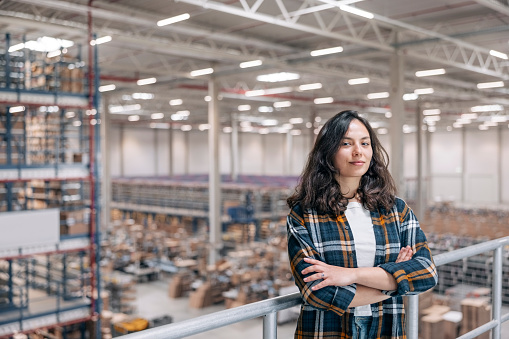 Portrait of a young woman working in a distribution warehouse. Female worker looking at camera with her arms crossed in the factory storage room.