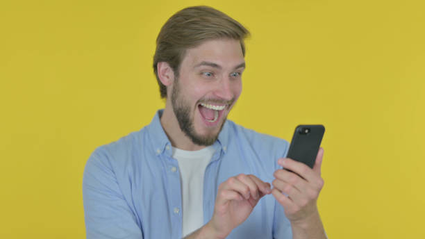 istock Young Man Celebrating on Smartphone on Yellow Background 1439570227