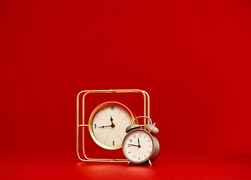 Gold clock next to a gray alarm clock against a red background with space for text