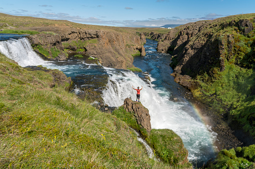 Woman with arms raised on rock enjoying Rejkjafoss waterfall on sunny day, North Iceland. achievement