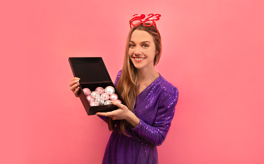 Happy smiling cheerful woman holding black box with Christmas balls, wearing purple sparkly glitter dress with red party glasses and looking at camera on a pink background.\n\nNew Year 2023 celebration.