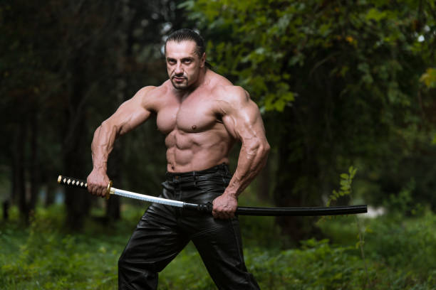 Action Hero Muscled Man Holding A Ancient Sword - Standing In Forest Wearing Leather Pants