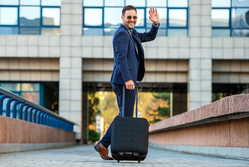 Elegante hombre de negocios con maleta saluda con la mano y va de viaje. Concepto de transporte photo