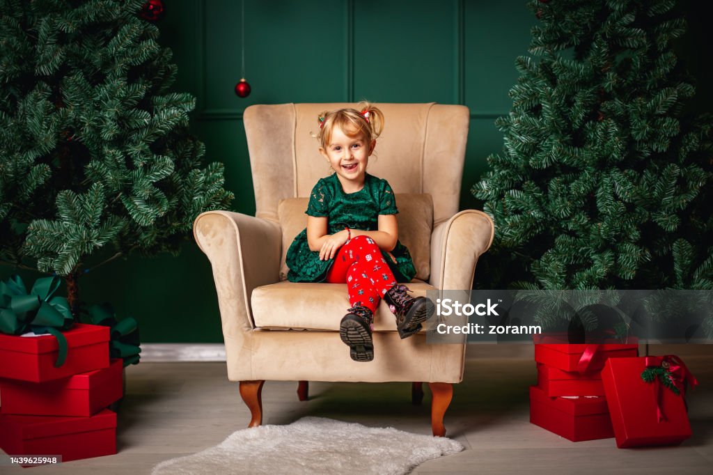 Cute little blond girl sitting in big armchair between Christmas trees with red gift boxes Cute little blond girl smiling happily at camera, sitting in large armchair between two Christmas trees with red gift boxes piled up Christmas Stock Photo