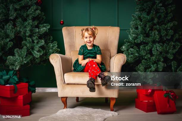 Cute Little Blond Girl Sitting In Big Armchair Between Christmas Trees With Red Gift Boxes Stock Photo - Download Image Now