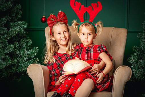 Cute blond sisters wearing bow and antlers headbands sitting together in armchair by Christmas tree, holding a book and smiling at camera