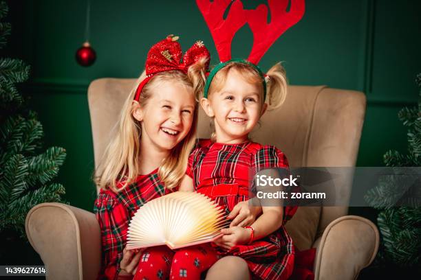 Two Beautiful Blond Girls Sitting Together In Armchair And Smiling With Joy In Christmas Décor Stock Photo - Download Image Now