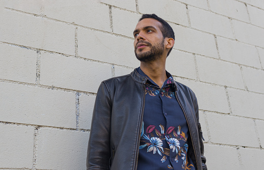 Portrait headshot of a homosexual pride guy dressed in flower shirt and leather jacket standing ahead a white brick white wall and looking something away