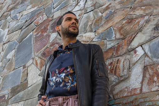 low angle waist-up shot of a hispanic gay guy dressed in flower shirt and leather jacket standing ahead a stone wall with one hand on hip while looking aside