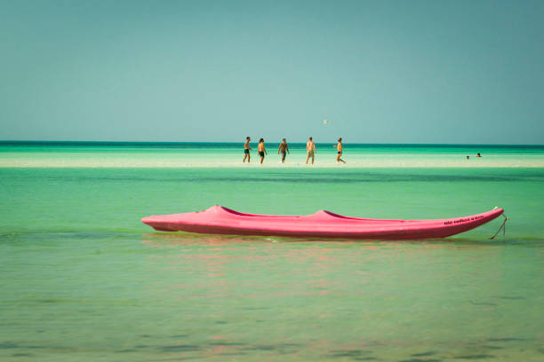 Isla Holbox, Yucatan, Mexico - April 11, 2021: Pink kayak floats in shallow water where five muscular male adults wearing swimming trunks are playing soccer on a sandbar in the Gulf of Mexico ocean.
