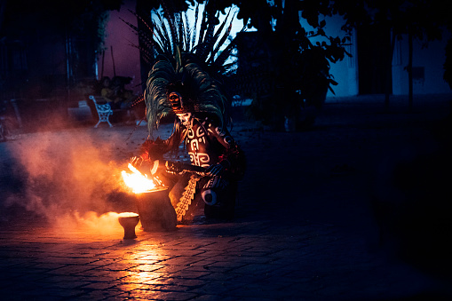 Yucatan, Mexico - April 15, 2021: An adult man dressed in a costume and larged feathered headdress with skeleton mask plays with fire at night in the village of Valladolid