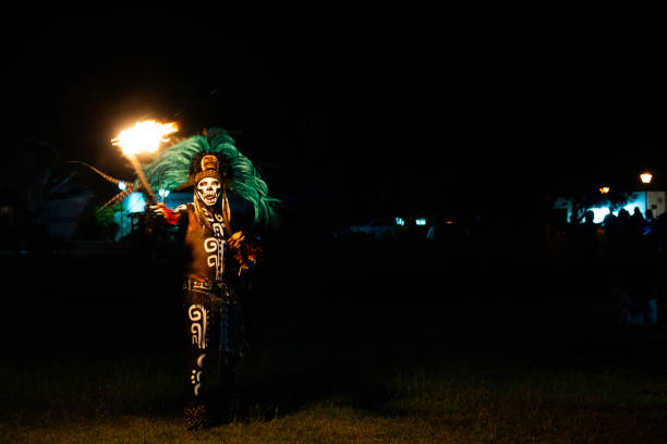 Yucatan, Mexico - April 15, 2021: A Hispanic adult man street performer dressed in a costume and larged feathered headdress with skeleton mask plays with fire at night in the village of Valladolid