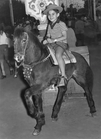 Black and white Image taken in the 50s: Smiling boy riding a small horse looking at the camera