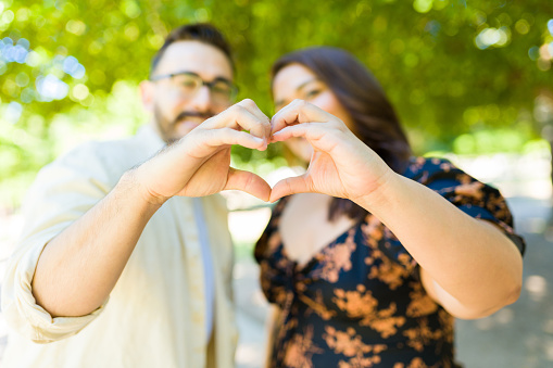 Focus on foreground of a loving young couple looking happy making a heart love gesture with their hands