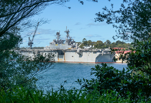 The USS Tripoli (LHA 7) of the US Navy docked at Garden Island, Sydney Harbour.  The red line in the water is a protective device around the ship.  This image was taken through the vegetation of Mrs Macquarie's Chair on an afternoon in Spring.