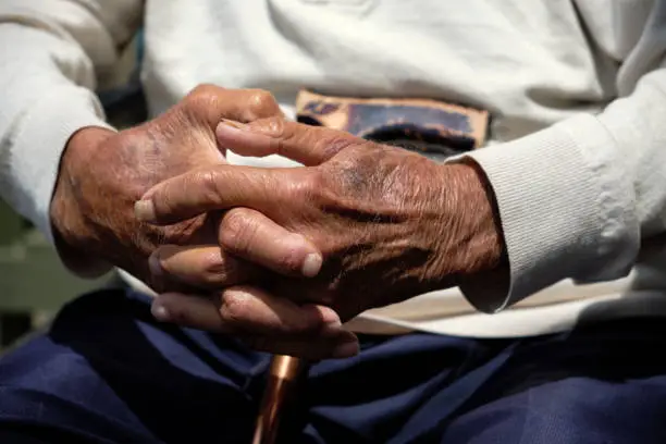 Dark skin toned man with hands clasped. Elderly man sitting with a walking stick with his hands clasped in his lap. Model wears light colored long sleeve jumper and dark blue trousers.