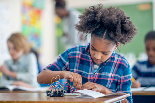 A young female student of African decent, sits at her desk as she diligently works away on a STEM project.  She is dressed casually and is focused on the task.