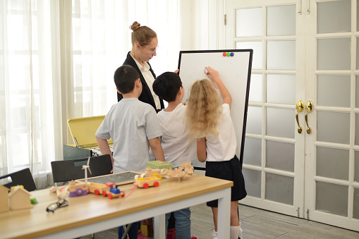 A mother or teacher is teaching her little students how to draw with colorful pens in the school classroom. The concept of parenting and home education By being creative and artistic children.