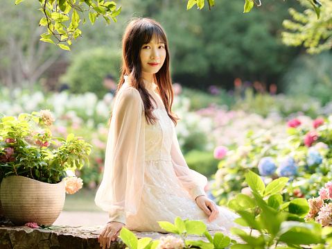 Beautiful woman in white dress posing in colorful hydrangea flowers field, backlit effect, charming Chinese girl with black long hair enjoy her leisure time outdoor.