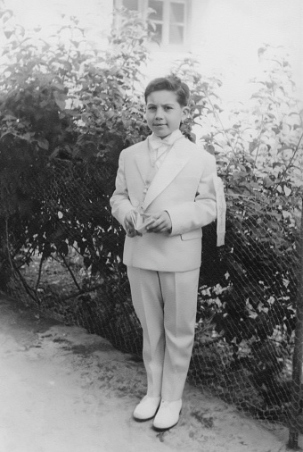 Black and white Image taken in the 50s: Smiling boy holding a missal posing with his first communion outfit looking at the camera