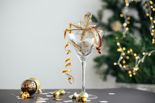 Merry Christmas and Happy New Year! Stylish christmas glitter star in champagne glass, golden confetti and baubles on background of festive lights. New year party creative concept. Moody banner