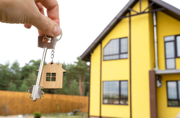 una mano con las llaves de una nueva casa en el fondo de una cabaña sin terminar. construcción, proyecto, mudanza a una nueva casa, hipoteca, alquiler y compra de bienes raíces. para abrir la puerta. espacio de copia - house real estate residential structure townhouse fotografías e imágenes de stock