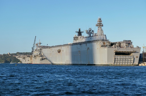 HMAS Canberra (L02) of the Royal Australian Navy and USS Tripoli (LHA 7) of the US Navy, docked at Garden Island, Sydney Harbour. \n The yellow buoy marks an exclusion zone around the US warship. A red coloured object floating around the ship operates as protection.  This image was taken at sunset in Spring, an office tower in the central business district casting a shadow over HMAS Canberra.