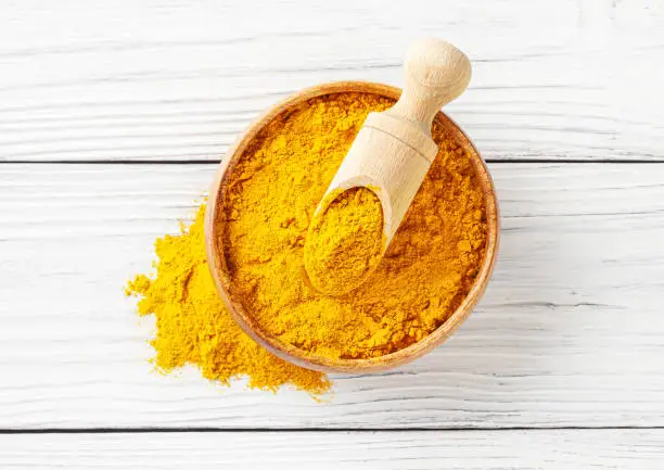 Dry turmeric (curcuma) powder in wooden bowl with scoop on white wooden background