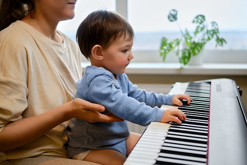 A mother woman teaches a toddler baby to play the piano in a home living room. An adult teacher with a child playing music on an electric piano. Kid boy age one year four months
