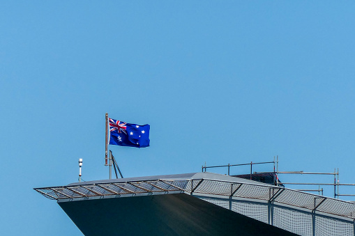The Australian flag flies on the flight deck of HMAS Canberra moored at Garden Island in Sydney Harbour.  Scaffolding surrounds the flight deck under repair.  This image was taken from Mrs Macquarie's Chair in Spring.