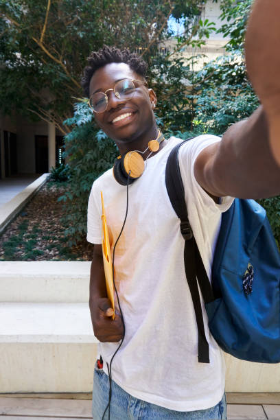 selfie vertical de um estudante africano usando fones de ouvido e segurando uma pasta de arquivos no campus. - male education college student nerd - fotografias e filmes do acervo