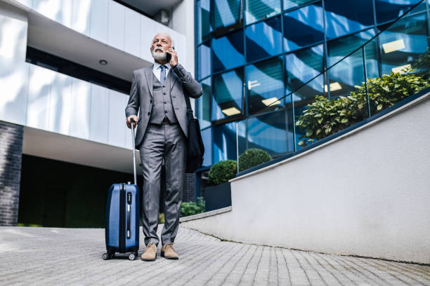 istock Serious elderly male professional with suitcase talking on smart phone while standing against office building. 1439695633