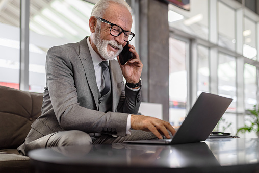 Confident senior businessman in suit using laptop while talking on smart phone at the office.