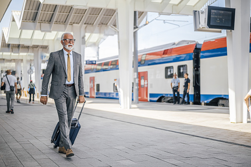 Elderly businessman in suit walking with luggage at subway or train railway station platform during sunny day.