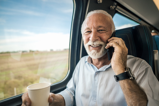 Smiling elderly professional businessman talking on mobile phone while having coffee by window in subway or train