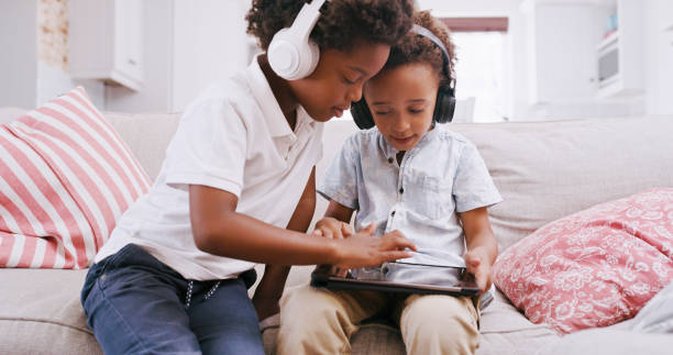 Headphones, tablet and children learning games in their home living room for education, digital development and holiday activity. Black family kids with audio, sound or music with a game app on sofa stock photo