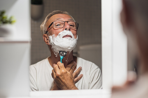 Senior man is shaving his beard with razor and shaving foam in bathroom.