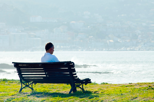 Nigrán,Spain- February 9, 2022: Man sitting alone on wooden bench in front of the sea. Playa América beach ,Nigrán, Rías Baixas, Pontevedra province,Galicia, Spain. Bayona townscape in the background.