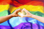 Two multiracial hands join together to form a shape heart with a LGBT flag background. Concept of homosexual gay pride day and love between different races, peace, humans against discrimination