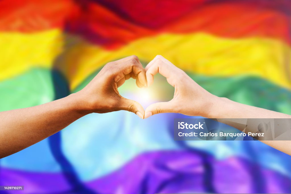 Two multiracial hands join together to form a shape heart with a LGBT flag background. Concept of homosexual gay pride day and love between different races, peace, humans against discrimination Two multiethnic hands join together to form a heart with a blue sky background. Concept of hope and love between different races, peace between humans and against hatred and racism. High quality photo LGBTQIA People Stock Photo