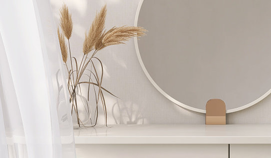 Minimal, modern and contemporary design of creamed colored dressing table with round vanity mirror, vase of pampas in dappled sunlight from window curtain in white wall bedroom for beauty and personal care product display